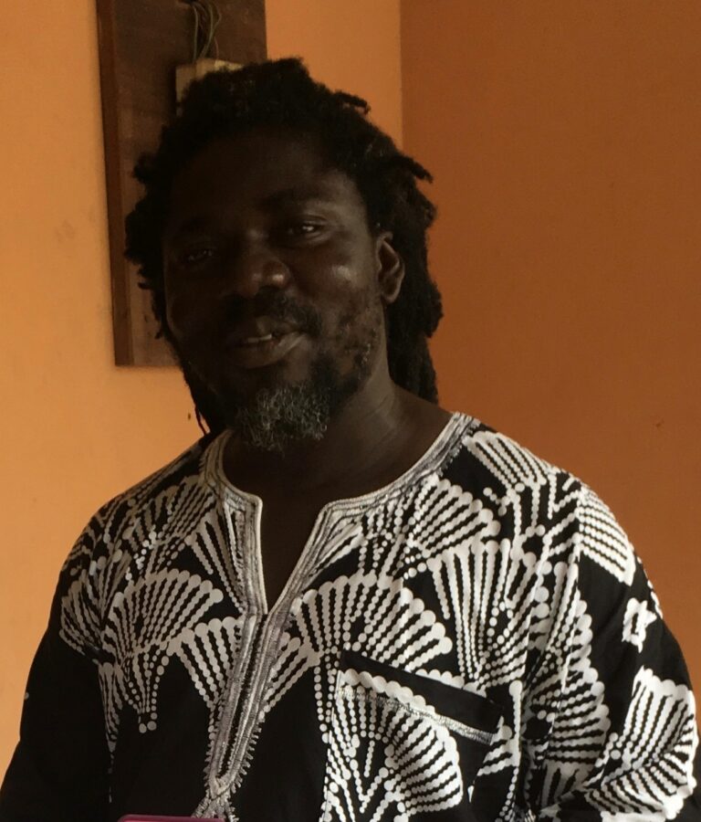 A man with dreadlocks standing in front of an orange wall.